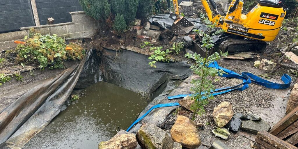 Blocked drain under pond being cleared with mini excavator and hoses for efficient water flow.